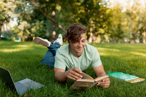 Cheerful caucasian student guy reading book outdoors, preparing for lectures while lying on lawn in university campus or park, free space