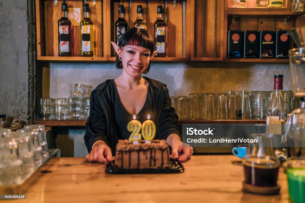 Woman barista standing behind a counter with a birthday cake Young smiling woman barista with elf ears standing behind a counter with a birthday cake Irish Culture Stock Photo