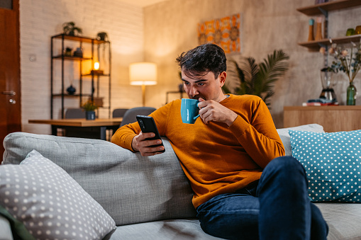 Smiling young handsome man relaxing at home, texting on smartphone and drinking coffee.