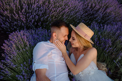 Young happy smiling couple, a woman in a white dress and hat and a man in a white shirt are lying down in a lavender field.