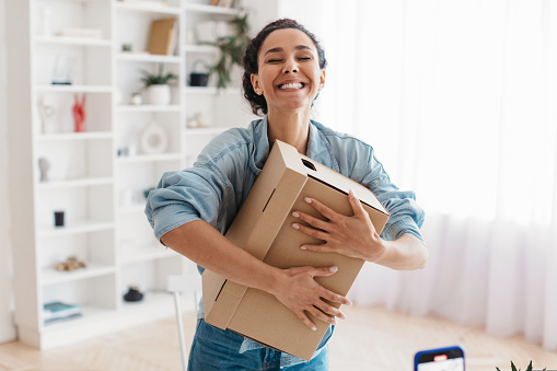 Shopping And Delivery Service. Joyful Middle Eastern Lady Holding Cardboard Box Receiving Delivered Package Standing At Home, Smiling With Eyes Closed. Shopaholism And E-Commerce Concept