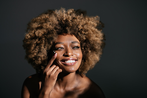 Studio shot of beautiful African-American woman with curly hair looking away and putting on moisturizing cream on her face.