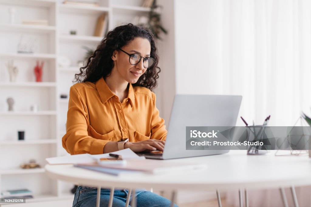 Middle Eastern Lady Using Laptop Working Online Sitting In Office Middle Eastern Lady Using Laptop Working Online Wearing Eyewear Sitting At Workplace In Modern Office. Remote Job, Technology And Career Profession Concept. Side View Women Stock Photo