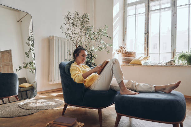 Attractive young woman reading a book while relaxing in a comfortable chair at home stock photo