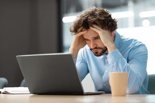 Error. Portrait of sad business man sitting at desk using pc, grabbing head looking at screen. Remote Work. Stressed guy suffering job problems, reading unexpected bad negative news, free copy space