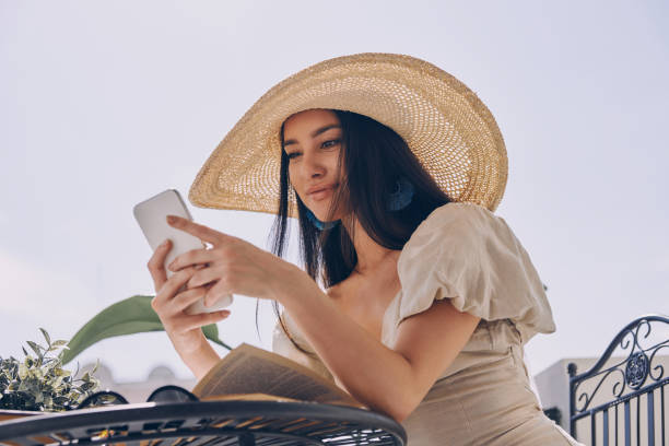istock Beautiful young woman in elegant hat using smart phone while relaxing on the balcony 1434215043