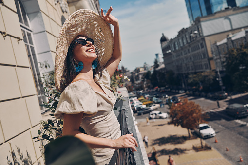 Relaxed young woman adjusting her elegant hat and smiling while standing on the balcony