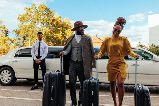 A wealthy African American couple is walking from the limousine to their hotel. They are carrying their luggage while the limo driver is standing next to the car in the background.