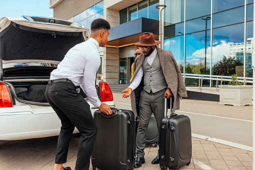 A mid adult African American couple arrives at their upscale resort for a luxurious weekend being helped by the limo driver with the luggage being taken out of the trunk.