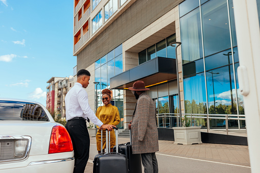 An African American couple is standing with their luggage next to the limo. They had just arrived to a five star hotel resort. They are on their weekend getaway. Driver is helping them with their suitcases.