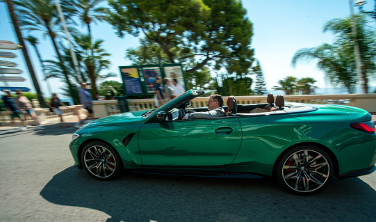 Amazing green convertible and a man in a jacket driving in Monaco and Palms and people walking
  on a beautiful day