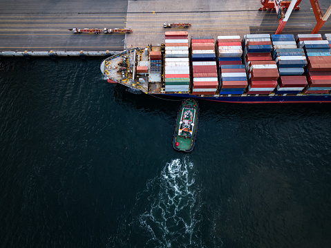 Aerial drone view of a container ship docking with the assistance of tugboats.