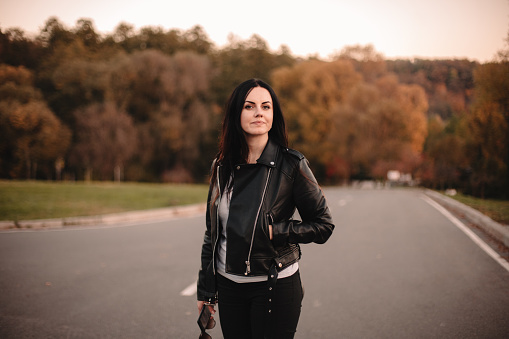 Portrait of happy stylish young woman wearing leather jacket standing on the road looking at camera during sunset in autumn