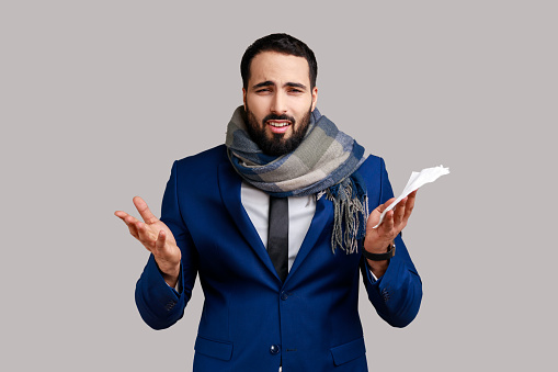 Bearded man wrapped in scarf holding handkerchief, shouting with hate anger, feeling annoyed from influenza disease, wearing official style suit. Indoor studio shot isolated on gray background.