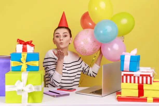 Photo of Woman with party cone sitting workplace surrounded by gift boxes and balloons, sending air kisses.