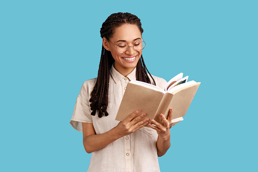 Smiling optimistic woman with black dreadlocks reading book and smiling, enjoying, standing absorbed with exciting plot, wearing white shirt. Indoor studio shot isolated on blue background.