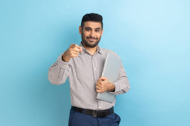 istock Businessman standing holding closed laptop or folder, has positive expression, pointing to camera. 1434251516