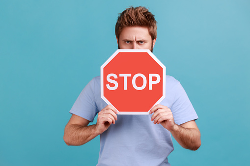 Portrait of serious strict bearded man covering half of face with red traffic sign, warning to go, prohibition concept, bossy expression. Indoor studio shot isolated on blue background.