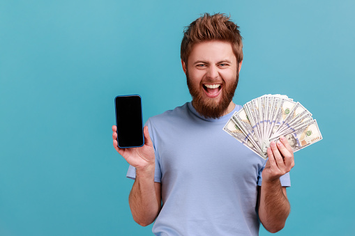 Portrait of extremely happy handsome bearded man holding dollar banknotes and cell phone with empty screen, online betting, winning. Indoor studio shot isolated on blue background.