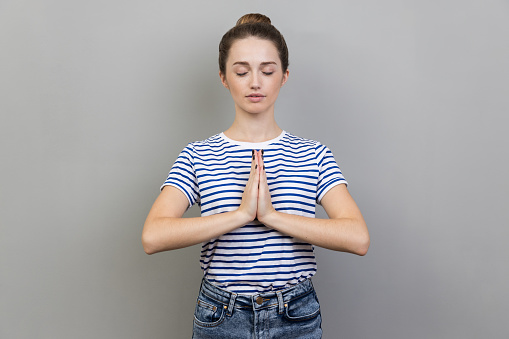 Portrait of woman wearing striped T-shirt keeps hands in yoga gesture, has calm facial expression, keeping palms pressed together and closed eyes. Indoor studio shot isolated on gray background.