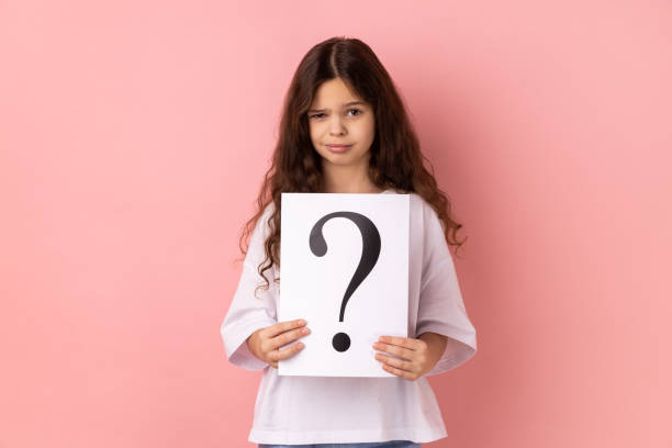 istock Winsome little girl looking at camera, holding paper with question mark, thinks about tasks. 1434251916