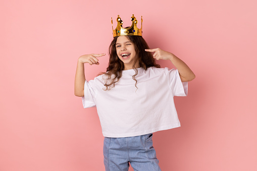 Portrait of happy confident smiling little girl wearing white T-shirt standing in gold crown, pointing at herself with pride, looking at camera. Indoor studio shot isolated on pink background.