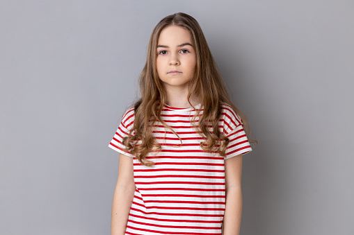 Portrait of strict serious little dark haired girl wearing striped T-shirt looking at camera with confident concentrated expression. Indoor studio shot isolated on gray background.