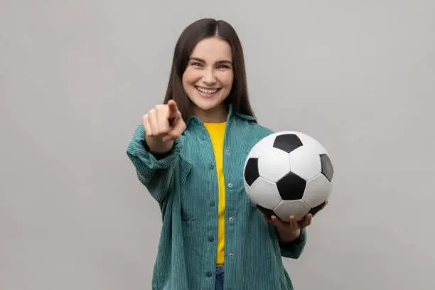 Photo of Beautiful woman holding soccer ball, looking at camera with toothy smile, pointing finger to camera.