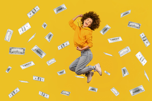 Portrait of successful rich woman with Afro hairstyle jumping in money rain, celebrating her victory, clenched fists and screaming happily. Indoor studio shot isolated on yellow background.