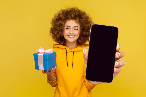 istock Cute delighted woman holding present box and showing big empty cell phone display for advertisement. 1434251990