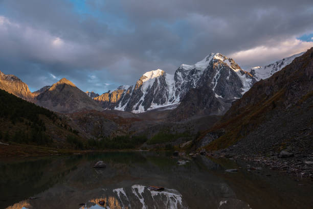 istock Dramatic landscape with giant snow mountains reflection in glacial lake. Beautiful mountain lake and sunlit gold snowy mountain top in cloudy sky. Huge snow-covered mountains in sunlight in overcast. 1434262875