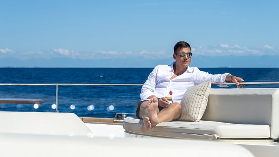 Successful man relaxing on sunny day on yacht while cruising on open sea. Sitting with drink on comfortable seat on deck. Luxury vacation at sea.
