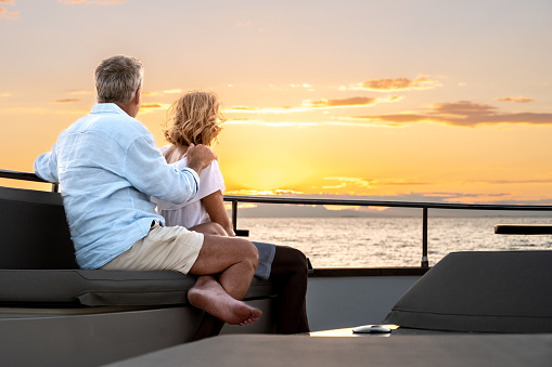 Back view of couple enjoying romantic sunset from yacht deck. Colourful sky with clouds. Luxury vacation at sea.