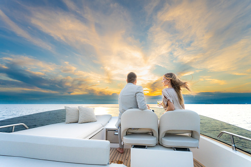 Back view of man and woman sitting on top deck of impressive yacht and watching romantic sunset. Luxury vacation at sea.
