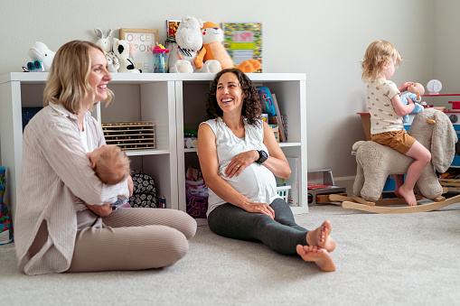 A mixed race pregnant woman enjoys talking with a friend who has a newborn baby and toddler daughter. The women are sitting on the floor of the older children's bedroom or playroom.