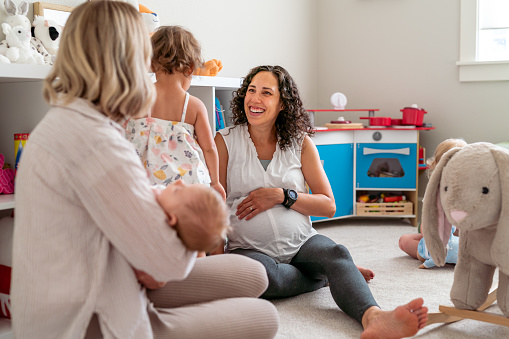 A mixed race pregnant mother sits on the floor at home and talks with her toddler daughter as the child has a playdate. A caucasian woman is also sitting on the floor, rocking her baby to sleep