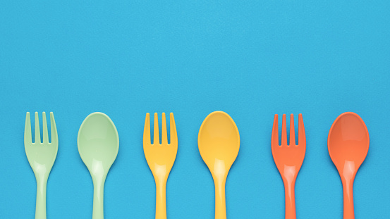 Orange, yellow and green plastic forks and spoons on a blue background. Minimal concept of plastic tableware. Flat lay.