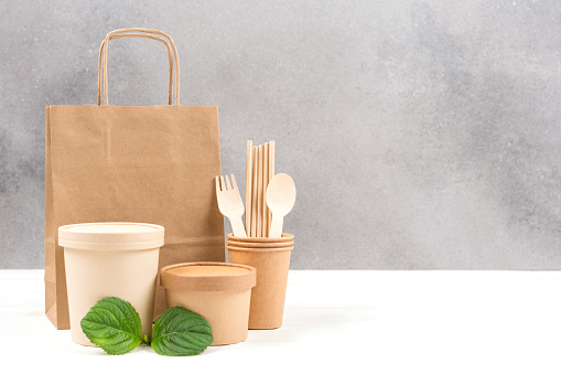 Paper utensils - food paper containers, bag, cups, drinking straws and wooden cutlery set against gray wall background with copy space. Sustainable food packaging. Eco tableware