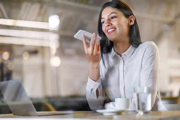 Happy businesswoman talking to virtual assistant on her phone while working in the office. The view is through glass.