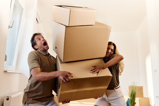 Mid adult couple cooperating while trying to carry heavy cardboard boxes in their new apartment.
