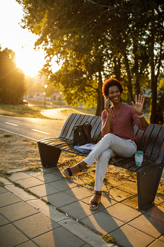 Full length shot of joyful mid adult woman sitting on the street bench, talking on the phone and waving at someone off camera.