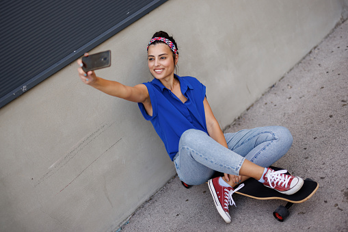 Smiling young woman sitting on her skateboard by the wall in the city and taking selfie on mobile phone