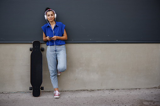 Young woman with skateboard leaning on gray wall and listening music on headphones in the city