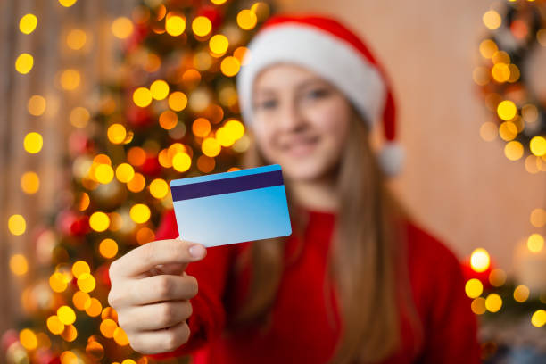 Hand of a girl, holding credit card close up on background of beautiful christmas tree with lights in festive room stock photo