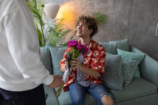 A young homosexual man surprising his boyfriend with a bouquet of flowers in their apartment living room