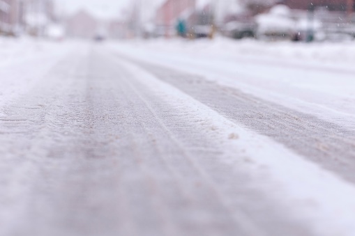 A low portrait of an asphalt road or street covered in snow and ice during winter. There are frozen tire tracks visible in the white surface, which makes it extra slippery and dangerous to drive on.
