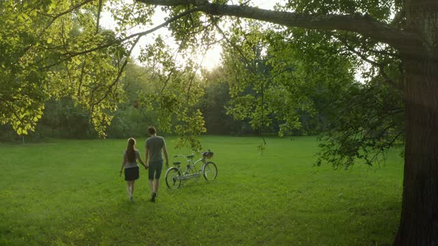 A couple walks across the grass to their tandem bike, hugs and kisses next to it