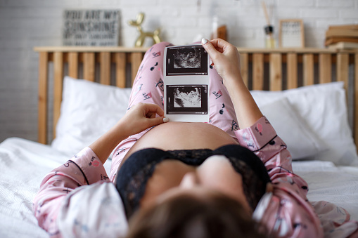 Partial nudity.A happy pregnant woman sitting on a white bed holds an ultrasound image in her hands,looks at the medical examination report of the unborn child.A pregnant female shows her ultrasound report and her unborn baby.The concept of pregnancy