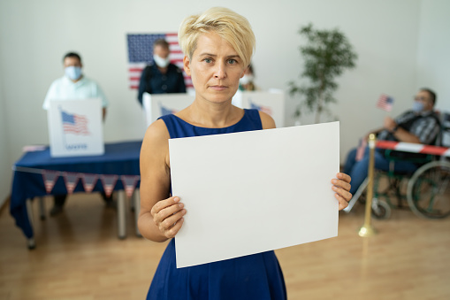 A mid-adult serious looking woman holding a blank sign is standing in a voting station with US flag in background