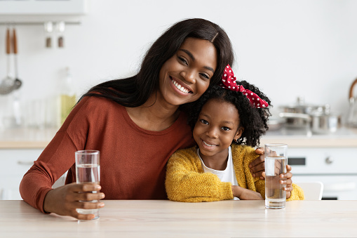 Happy African American Family Mother And Daughter Holding Glasses With Water While Sitting Together At Table In Kitchen, Smiling Black Mom And Female Child Enjoying Healthy Drink, Free Space